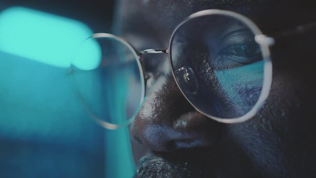 Close up tilt up shot of face of African American programmer in glasses working in dark room with blue neon light