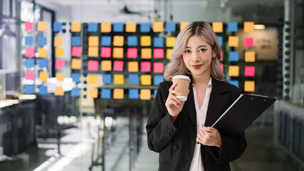 Charming Asian woman with a smile standing holding papers and coffee cup at the office.
