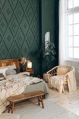 Bedroom interior with emerald walls and a large window, a large wooden double bed with pillows and blankets, a wooden bench and a large wicker chair near the window. Plants in pots. Empty space