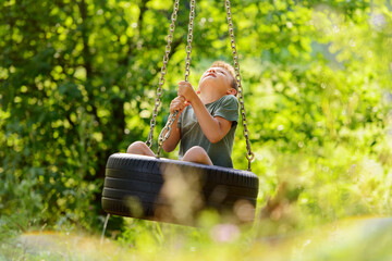 Active boy on a colorful homemade swing tire in the forest. Healthy summer activity for kids in sunny weather. Forest playground.