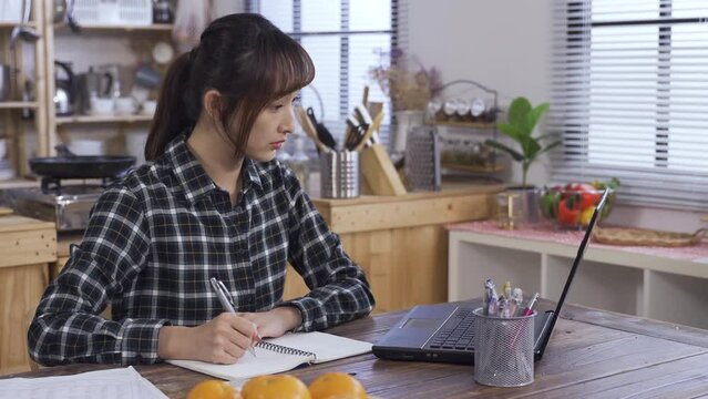 asian young female is making notes and typing on the laptop while elearning at dining table from home.