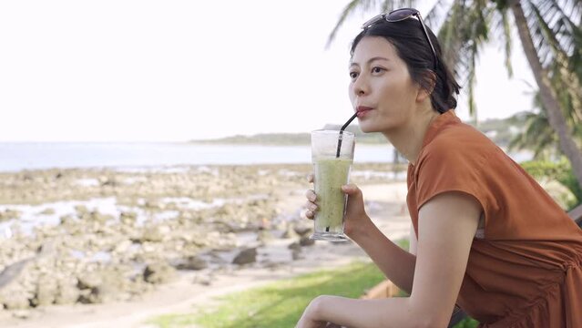 happy asian female is gazing into distance while relaxing by the deck railing to enjoying her fruit smoothie and the beautiful beach on a clear day.
