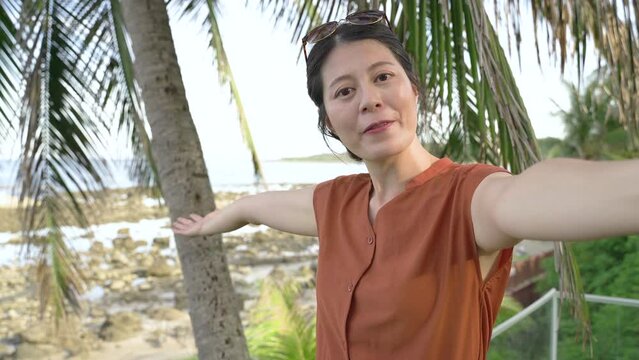happy asian influencer filming vlog at a beach resort near a palm tree is waving hi, gesturing to the open beach while introducing and saying bye to the camera