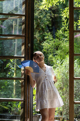 Young woman stands leaning on French window frame and holds fan blowing because of hot day. Blonde lady shakes fan to cool off suffering of hot tropical weather against exotic garden outside closeup