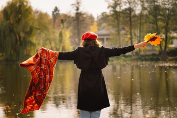 A Teengirl in a red beret with a bouquet of autumn leaves in her hands walks on wooden pontoon. Autumn mood. Autumn concept. Autumn day. Back view