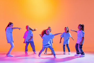 Naklejka premium Group of children, little girls in sportive casual style clothes dancing in choreography class isolated on orange background in purple neon light.
