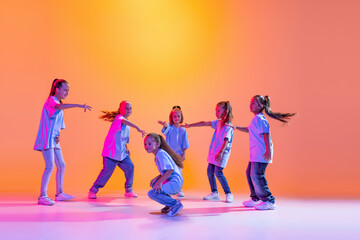 Portrait of cheerful, active little girls, happy kids dancing isolated on orange background in neon...