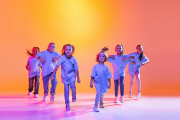 Group of children, little girls in sportive casual style clothes dancing in choreography class isolated on orange background in purple neon light. - obrazy, fototapety, plakaty