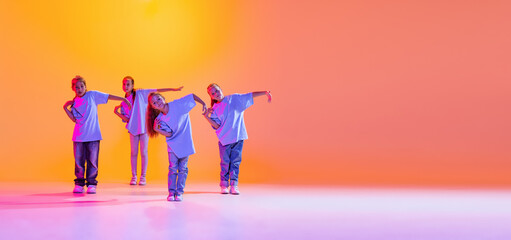 Dance group of happy, active little girls in t-shirts and jeans in action isolated on orange background in neon. Concept of music, fashion, art, childhood