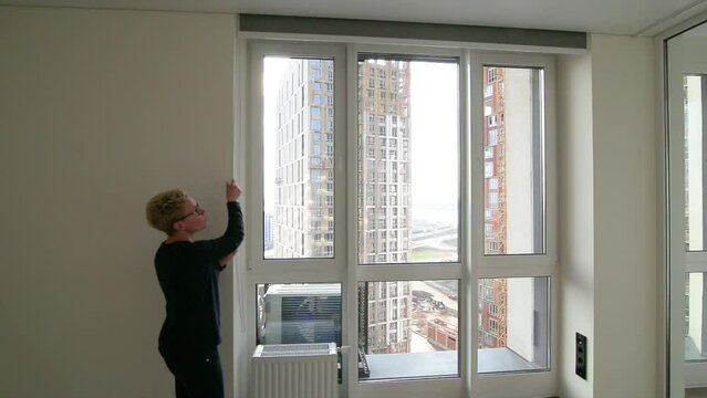 The girl lowers the roller shade on the large window. Gray fabric. Outside the window is the city, skyscrapers.