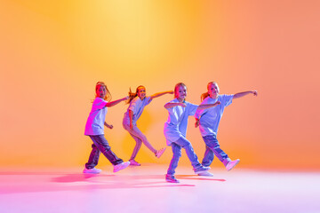 Dance group of happy, active little girls in t-shirts and jeans in action isolated on orange background in neon. Concept of music, fashion, art, childhood