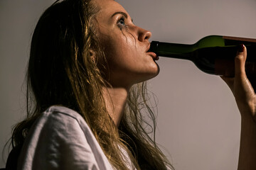 Crying young woman with smeared mascara drinks alcohol from neck of bottle. Side view portrait of...