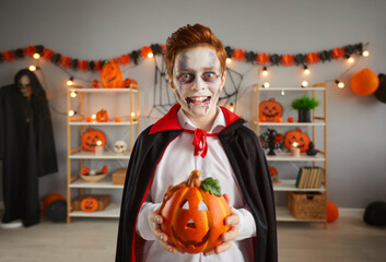 Kid in spooky Halloween vampire costume. Child dressed up as Count Dracula holding jack-o-lantern decor. Portrait of boy in black cape with crazy eyes, excited expression and fake blood makeup on face - obrazy, fototapety, plakaty