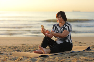 mature woman takes a break while sitting and relaxing on the yoga fitness mat and using smartphone on the beach by the sea in the morning. Woman outdoor workout. Healthy, chill out lifestyle.