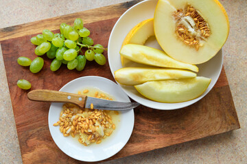 sliced melon with seeds on a white plate, a kitchen knife and green grapes on a wooden board, the concept of fresh fruit and healthy eating