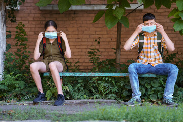 schoolchildren a boy and a teenage girl are sitting on a bench outside the school building, they use a protective breathing mask against covid or other respiratory infection