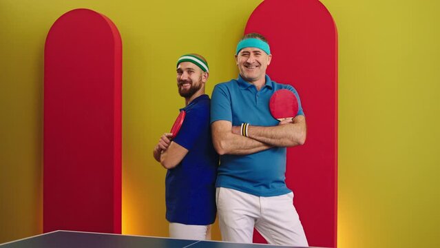 Excited father and son posing in front of the camera with the ping pong racket after they end the game looking straight to the camera