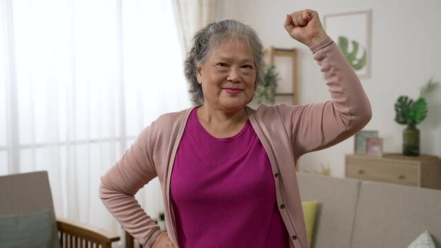 portrait of a confident asian grandmother lifting her arm in posture of power and smiling at the camera in the living room at home
