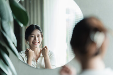 A young Asian woman talks to herself through a mirror to build her self-confidence and empower...