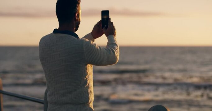 Man filming and taking photos of beach after sunset on phone while on vacation. Taking pictures of holiday by the ocean in the evening. Happy male tourist making a video on the coast with smartphone.