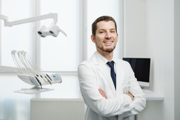 Fototapeta premium Happy male dentist in white lab coat posing with hands crossed and looking at camera