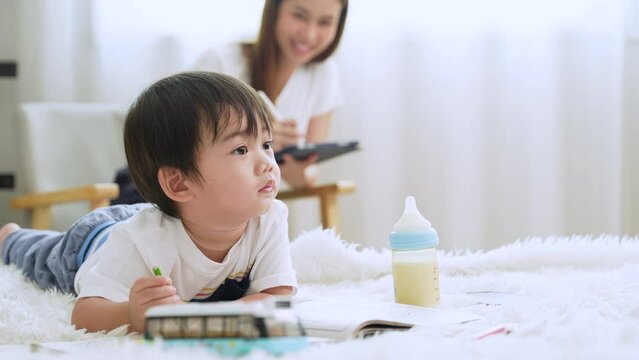 Cute little Asian boy playing painting on the bed in the bedroom with his mother sitting and working not far away.