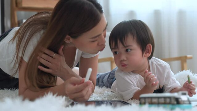 Asian young mother kisses her little son and take the little son to paint to promote development in the living room at house.