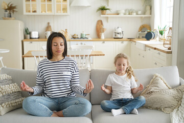 Mother, foster daughter practice yoga sitting on sofa at home. Healthy lifestyle, adoption of child
