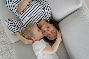 Fototapeta na wymiar Little daughter kiss smiling mum on cheek, relaxing on couch at home. Tender moment of motherhood