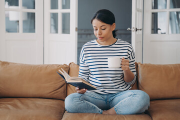 Obraz premium Peaceful young caucasian woman is reading book sitting on leather couch. Stress relief concept.