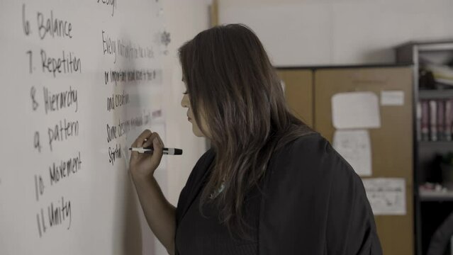 Native American teacher writes the principles of graphic design on a white board and erases a mistake with her finger.