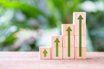 Wooden cubes stacked as step staircase with increasing green arrow symbol arranged on the table with green plant on the background, green business trend and growth, career progress concept