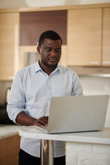 Serious Black man standing at kitchen counter and checking working chats on laptop