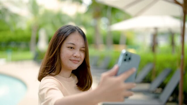 4K, A close-up shot of a young Asian tourist taking a selfie with her cell phone happily at the poolside of her hotel on a weekend.