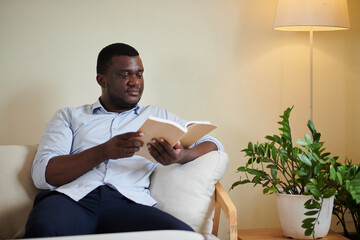 Portrait of Black man sitting on comfy couch at home and reading captivating book