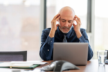 Stressed middle aged businessman having a migraine while sitting at office desk and working on laptop