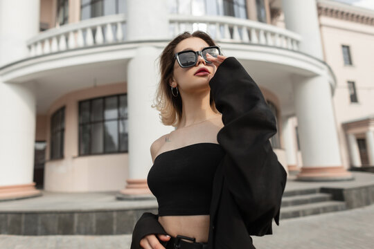 Fashion elegant pretty woman model in stylish trendy black clothes with top and blazer wears a cool modern sunglasses and walks in the city. Urban female business style look outfit