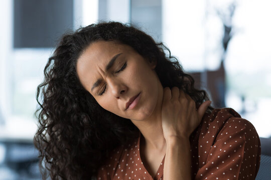Close-up photo of curly-haired woman at home near the window with severe neck pain, business woman massaging her neck with closed eyes
