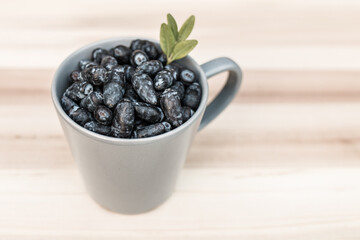 Kamchatka berry / haskap berry / honeysuckle (Lonicera caerulea) in a gray cup, decorated with green leaves, standing on a wooden board. The first summer fruit, perfect for desserts. - obrazy, fototapety, plakaty