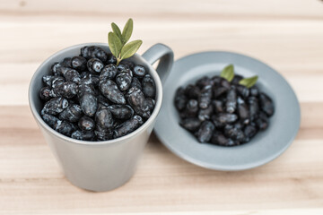 Kamchatka berries / haskap berries / honeysuckle (Lonicera caerulea) in a gray cup and on a gray plate decorated with leaves, placed on a wooden board, side view. - obrazy, fototapety, plakaty