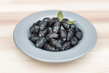 Freshly picked haskap berries / honeysuckle (Lonicera caerulea) on a gray plate, on a wooden board, with green leaves. Organic healthy berries full of vitamins. - obrazy, fototapety, plakaty