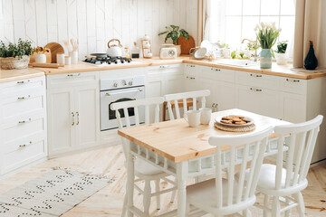 Wooden dining table with white chairs on a blurred background of cabinets in a Scandinavian modern kitchen interior filled with kitchen accessories. White kitchen with appliances and large windows