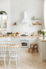 Modern white Scandinavian style kitchen with wooden worktop filled with kitchen accessories. Stylish kitchen interior with dinner table, cooker hood, built-in oven, gas hob and large windows. Nobody