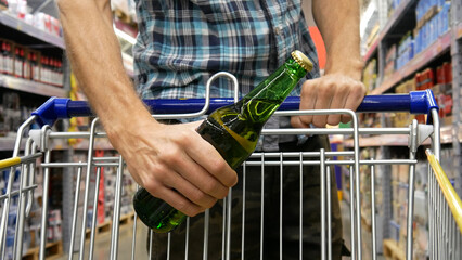 Close-up of a shopping trolley in a supermarket and a man puts a green glass bottle of beer into it