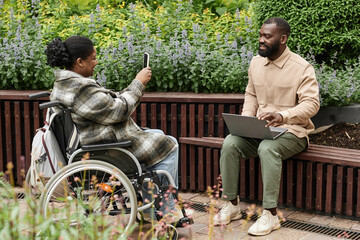 Portrait of smiling black woman in wheelchair taking mobile photo of friend or partner outdoors, copy space