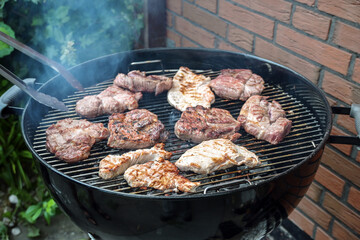 Steaks of various meats on a mobile round grill are roasted over charcoal at a barbecue party in the backyard, outdoor cooking concept, copy space, selected focus
