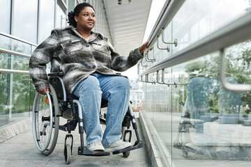 Full length portrait of smiling black woman in wheelchair moving down ramp in city, urban...