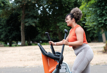 Fototapeta na wymiar Smiling chubby young mixed race woman exercising on elliptical trainer in a London Park.