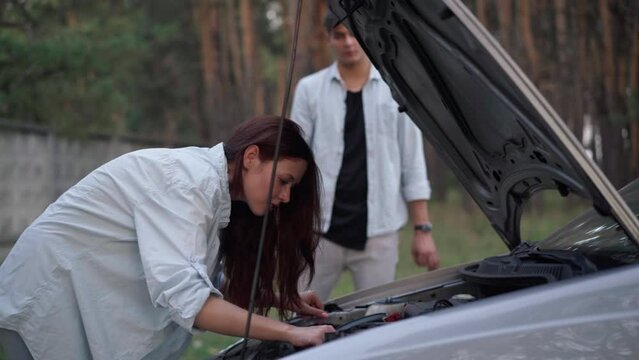Side view confident young woman repairing automobile leaning at open car hood with blurred man talking at background. Concentrated Caucasian girlfriend with anxious boyfriend in forest