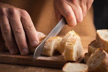 Whole grain bread close-up on the wooden cutting board. Man cutting fresh bread with a big knife. Knife with serrated blade. Fresh bread on the kitchen table. Traditional pastries in bakery
